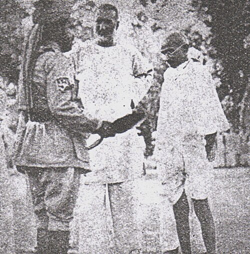 Heavily degraded black and white photograph of three or four figures standing outdoors, one in a uniform with an armband, the others in white.