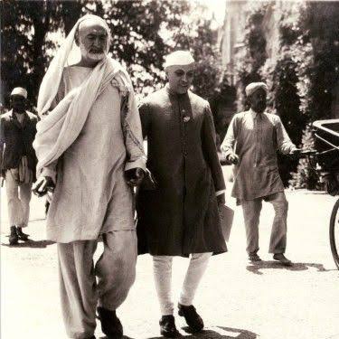 Black and white photograph: Ghaffar Khan, tall and wrapped in a shawl, walking along a road beside Jawaharlal Nehru, with other figures and a horse-drawn carriage behind.