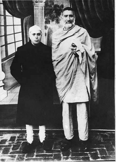 Formal black and white photograph: a young Jawaharlal Nehru in a dark coat and white cap standing beside a much taller Ghaffar Khan wrapped in a pale shawl, on a brick-paved floor.