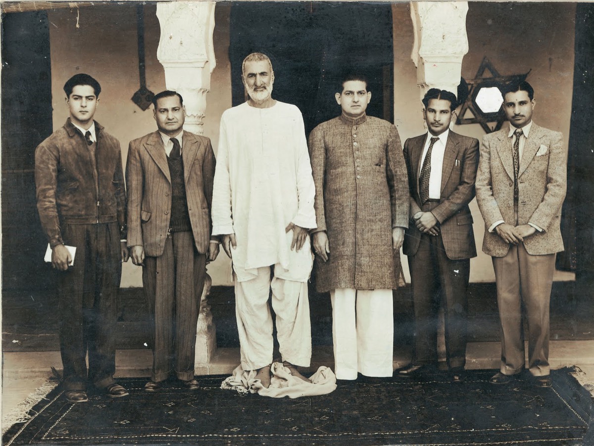 Sepia group photograph: Khan Abdul Ghaffar Khan stands at the centre, tall, barefoot, in plain white homespun, flanked by six men in suits and a sherwani, on a patterned carpet before an arched colonnade.