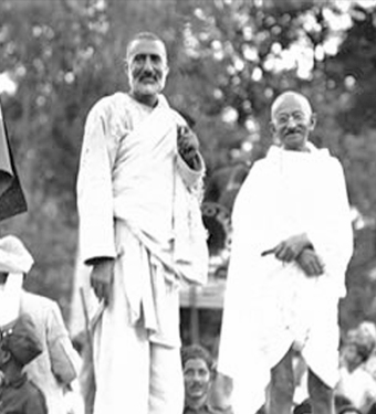 Black and white photograph of a public gathering: Ghaffar Khan standing at full height in white, with Mahatma Gandhi beside him in a shawl, surrounded by a crowd.